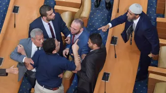 L'image montre une scène tendue dans une assemblée ou un parlement. Plusieurs hommes discutent vivement, certains pointant du doigt et affichant des expressions agitées. Il y a une mise en scène de désaccord, avec des gestes éloquents et une proximité physique entre les participants. Le décor est simple, avec des sièges en bois et un tapis bleu au sol, suggérant un environnement formel.