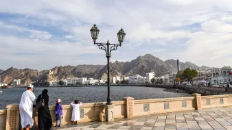 L'image montre une scène de bord de mer, probablement à Muscat, Oman. Dans le premier plan, on peut voir une famille vêtue de vêtements traditionnels. Un homme et une femme se tiennent près d'un lampadaire, regardant vers l'horizon. À côté d'eux, deux enfants, dont un porte une casquette rouge. En arrière-plan, on aperçoit des montagnes majestueuses ainsi que des bâtiments blancs typiques de la région. Le ciel est partiellement nuageux, créant une ambiance paisible et contemplative.