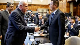L'image montre une scène de réunion ou de conférence, probablement dans un cadre formel. Deux hommes se serrent la main, souriant, ce qui évoque une interaction chaleureuse ou un accord. En arrière-plan, on aperçoit d'autres participants qui semblent également engagés dans des discussions. La salle est spacieuse, avec une décoration élégante, des fenêtres laissant passer une lumière naturelle, et des sièges disposés autour d'une grande table. L'atmosphère semble professionnelle et collaborative.