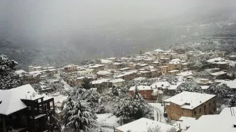 L'image montre un paysage hivernal où une petite ville est recouverte de neige. Les toits des maisons, en grande partie en bois, sont également blancs, créant une atmosphère paisible et froide. On peut apercevoir des arbres chargés de neige et des collines en arrière-plan, le tout enveloppé par une brume légère, ajoutant à la sérénité de la scène. La vue donne une impression de calme et d'isolement, typique des régions montagneuses en hiver.