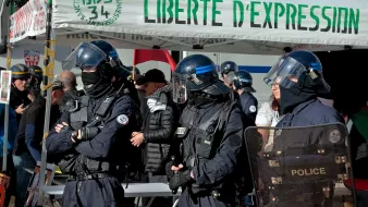 L'image montre un groupe de policiers en uniforme, équipés de casques et de protections, se tenant devant un stand ou une tente avec l'inscription "LIBERTE D'EXPRESSION". On peut voir plusieurs personnes en arrière-plan, probablement des manifestants ou des observateurs, dans un environnement urbain. L'atmosphère semble être celle d'un rassemblement ou d'une manifestation, avec des thèmes liés à la liberté d'expression.