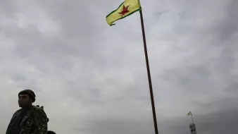 L'image montre un homme en uniforme militaire se tenant près d'un drapeau. Le drapeau est de couleur jaune avec une étoile rouge au centre, entouré de bandes vertes. L'arrière-plan est nuageux, ce qui donne un ton sombre à la scène. En plus de l'homme, on peut apercevoir une autre instance du drapeau en arrière-plan, également hissé sur un mât.