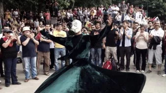 L'image montre une scène de rassemblement en plein air où une personne vêtue d'une longue robe vert foncé tourne gracieusement. Cette personne porte un masque à gaz, ce qui ajoute une dimension étrange et intrigante à la performance. Autour, une foule observe, certains prenant des photos avec leurs téléphones. L'ambiance semble à la fois festive et solennelle, avec des expressions variées parmi les spectateurs. Les arbres en arrière-plan ajoutent une touche de nature à la scène.