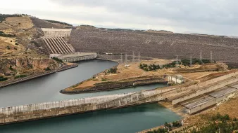 L'image montre une vue panoramique d'un complexe de barrages ou d'une centrale hydroélectrique. On peut voir un grand réservoir d'eau entouré par des collines sur lesquelles se trouvent des structures en béton. Des canaux ou des tunnels d'acheminement de l'eau sont visibles, ainsi que des installations de contrôle des niveaux d'eau. Le paysage est principalement aride, avec quelques zones verdoyantes et une atmosphère dramatique due à un ciel nuageux. Les infrastructures semblent bien entretenues et sont probablement liées à la production d'énergie.