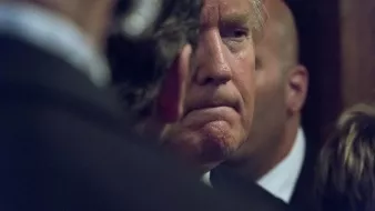 L'image montre un homme avec des cheveux blonds, affichant une expression pensive ou sérieuse. Il est partiellement obscurci par des personnes en arrière-plan, ce qui crée une atmosphère de concentration ou d'intensité. On peut également remarquer des costumes sombres, suggérant un cadre formel.