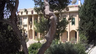 L'image montre un bâtiment en pierre, typique de l'architecture méditerranéenne, avec plusieurs arches et fenêtres. Il est entouré de verdure, avec des arbres et des plantes bien entretenues. Au fond, on remarque un satellite et un ciel bleu clair, ce qui suggère un climat ensoleillé. L'ensemble dégage une atmosphère paisible et accueillante.