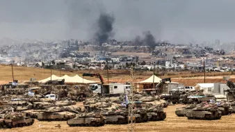 Un champ militaire avec des chars, fumée au loin et destructions visibles en arrière-plan.