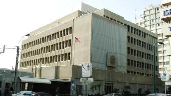 L'image montre un bâtiment moderne avec une architecture fonctionnelle. Il est en béton, avec plusieurs fenêtres rectangulaires sur la façade. Un drapeau américain est visible sur le toit, indiquant que c'est une représentation diplomatique. À l'avant du bâtiment, il y a des panneaux de signalisation et quelques véhicules garés le long de la route. L'environnement semble urbain, avec d'autres immeubles en arrière-plan.