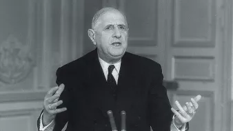 L'image montre un homme en costume formel, assis à une table avec un microphone devant lui. Il semble être en train de parler ou de faire un discours, utilisant des gestes expressifs avec ses mains. L'arrière-plan est sobre, avec des murs ornés de motifs simples. L'éclairage met en valeur son visage, soulignant son sérieux et son engagement dans son discours.