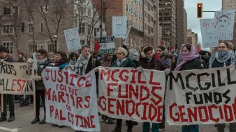 L'image montre une manifestation dans une ville urbaine. Un groupe de personnes est rassemblé, tenant des pancartes avec des messages en faveur de la Palestine et s'opposant au financement de ce qu'ils appellent un génocide. Certains manifestants portent des écharpes qui semblent indiquer leur solidarité avec la cause palestinienne. L'atmosphère est engagée et déterminée, avec des passants et des bâtiments en arrière-plan.