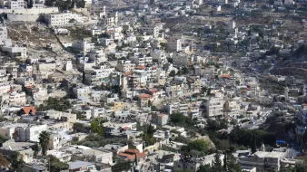 L'image montre un paysage urbain densément construit avec de nombreuses maisons et bâtiments. On peut observer une variété d'architecture, allant des structures modernes aux constructions plus anciennes. Les collines environnantes sont également visibles, avec des zones verdoyantes et des routes serpentant à travers le paysage. La ville semble être située dans une région montagneuse, avec un éclairage naturel qui met en valeur les détails des bâtiments.