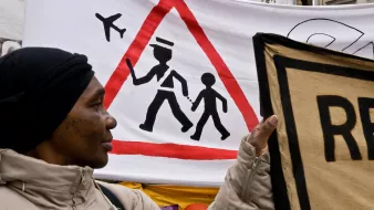 L'image montre une manifestation avec une femme tenant une pancarte. En arrière-plan, il y a une grande bannière avec un symbole d'avertissement en forme de triangle, représentant un adulte marchant avec un enfant. Ce symbole est accompagné d'illustrations d'avions en haut, suggérant une thématique liée au transport ou à la sécurité. La femme porte un manteau clair et un foulard noir, et son expression pourrait refléter l'engagement dans la cause qu'elle soutient.