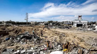L'image montre un paysage dévasté, probablement dans une zone de conflit ou après une catastrophe. Le sol est jonché de débris, avec des morceaux de béton et de métal éparpillés. On peut apercevoir quelques personnes se déplaçant parmi les ruines, tandis qu'à l'arrière-plan, des bâtiments endommagés se dressent sous un ciel partiellement nuageux. Les lignes électriques sont visibles, indiquant une infrastructure perturbée. L'ambiance apparaît sombre et désolée, témoignant de la destruction qui a eu lieu.