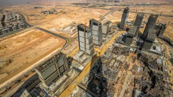 L'image montre une vue aérienne d'une région urbaine en développement, probablement dans un environnement désertique. On peut voir plusieurs gratte-ciels en construction, avec des structures modernes et des tours au milieu. Le paysage environnant est principalement désertique, avec peu de verdure, et des routes qui serpentent à travers la zone. La perspective donne une impression de grandeur et de modernité, tout en mettant en évidence les travaux d'infrastructure en cours.