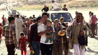 L'image montre un grand groupe de personnes se déplaçant, probablement dans un contexte de crise. On voit des adultes et des enfants marchant, avec des expressions sérieuses sur leurs visages. Certains portent des objets, et il y a une camionnette dans le fond. L'environnement semble désolé, avec peu d'infrastructures visibles. Cette scène évoque des thèmes de déplacement et de lutte, typiques de situations de conflit ou de catastrophe.