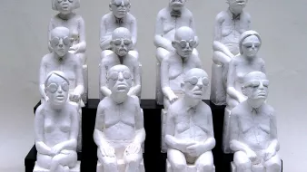 L'image représente une sculpture où plusieurs figures humaines, toutes de couleur blanche, sont assises sur des bancs. Les personnages sont stylisés avec des traits simples et géométriques, portant des lunettes, et semblent avoir des expressions neutres ou pensive. Ils sont agencés en un groupe de dix, disposés en plusieurs rangées, créant une impression d'unité et de contemplation. Les sculptures semblent évoquer des thèmes tels que la solitude, la réflexion ou la condition humaine.