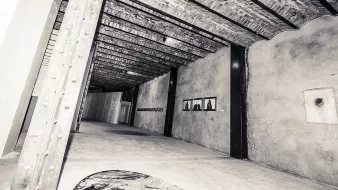 L'image présente l'intérieur d'un espace d'exposition ou d'une galerie d'art, avec de hauts plafonds en poutres de bois. Les murs sont en béton brut, créant une ambiance industrielle. Sur le mur de droite, on peut apercevoir quelques cadres accrochés, probablement contenant des œuvres d'art. Le sol est neutre, avec une texture qui semble lisse. L'éclairage met en valeur les détails architecturaux de l'intérieur, tandis que l'ensemble dégage une atmosphère contemporaine et artistique.