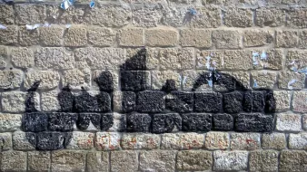 L'image montre un mur de pierres avec un graffiti en forme de silhouette noire. Le mur semble ancien et légèrement abîmé, avec des morceaux de papier déchirés. La silhouette créée par le graffiti évoque des formes humaines, mais sans détails précis. Les pierres du mur sont bien visibles et ajoutent une texture rustique à l'ensemble.