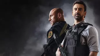 L'image montre deux hommes en tenue tactique, se tenant dos à dos dans une ambiance sombre et intense. L'un d'eux porte un uniforme de police ou militaire, tandis que l'autre est habillé d'une chemise et d'un gilet pare-balles. Ils semblent prêts pour une situation d'urgence, chacun tenant une arme à feu. Le fond est flou et dramatique, ajoutant à la tension de la scène.