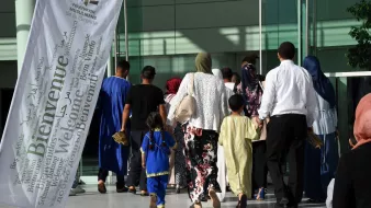 L'image montre un groupe de personnes se dirigeant vers un bâtiment. On peut voir des individus de différentes générations, y compris des enfants. Certains portent des vêtements traditionnels, et plusieurs femmes ont des foulards. Il y a également une grande bannière blanche flottant au vent, portant le mot "Bienvenue" en plusieurs langues. L'ambiance semble chaleureuse et accueillante.