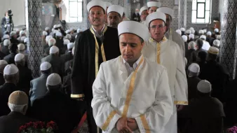 L'image montre un groupe d'hommes portant des vêtements traditionnels, notamment des robes blanches et des turbans. Ils semblent être en procession, probablement lors d'un événement religieux. En arrière-plan, de nombreuses personnes sont assises, probablement en prière ou en attente d'une cérémonie. L'architecture de la pièce inclut des motifs décoratifs et des fenêtres qui laissent entrer la lumière, créant une atmosphère solennelle.