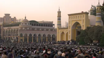 L'image montre une grande foule de personnes rassemblées dans une zone en plein air, probablement pour un service religieux. À l'arrière-plan, on aperçoit des bâtiments architecturaux impressionnants, avec des minarets et des ornements qui évoquent une ambiance culturelle riche. Les gens sont principalement assis, attentifs à ce qui se passe, créant une atmosphère de commémoration ou de prière collective. La lumière du soir donne une teinte chaleureuse à la scène.