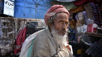 L'image montre un homme âgé avec une longue barbe grise, portant un turban rouge et blanc. Il semble regarder fixement avec une expression pensante ou préoccupée. L'arrière-plan suggère un marché ou un bazar, avec des étals et des objets divers. L'éclairage est naturel, ce qui donne une atmosphère vivante à la scène. L'homme porte un sac, ce qui indique qu'il pourrait être en train de se déplacer ou de faire des courses.