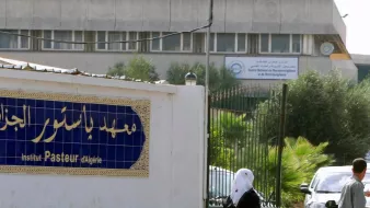 L'image montre l'extérieur de l'Institut Pasteur en Algérie. On peut voir un bâtiment moderne en arrière-plan avec des panneaux d'affichage. À l'avant-plan, il y a une entrée avec une façade blanche où figure le nom de l'institut en caractères arabes et français. Quelques personnes marchent dans cette zone, suggérant un environnement fréquenté. Des arbres et de la végétation ajoutent une touche de verdure au décor.