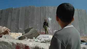 L'image montre un jeune garçon observant un soldat qui se tient devant un mur en béton. Le mur est marqué par des graffitis, suggérant un environnement de conflit ou de tension. Des débris sont dispersés au sol, et le ciel est dégagé, ce qui contraste avec la gravité de la scène. Le regard du garçon semble interrogateur ou curieux face à la situation qui l'entoure.
