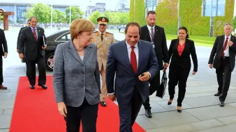 L'image montre un moment officiel, probablement lors d'une rencontre diplomatique. Une femme, portant un blazer gris, marche sur un tapis rouge aux côtés d'un homme en costume sombre. Ils semblent engagés dans une conversation. À l'arrière-plan, on peut voir des personnes en costume, ainsi que des bâtiments modernes et une végétation. L'atmosphère est formelle et professionnelle.