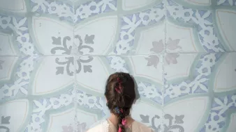 Cette image montre une personne de dos, portant un vêtement léger et délicat. Sa longue tresse, ornée de fleurs colorées, est visible et se détache sur un fond de motifs bleu clair. Les motifs sont ornés de formes géométriques et florales, créant une ambiance apaisante et artistique. L'ensemble évoque une atmosphère de calme et de beauté.