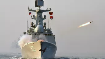 L'image montre un navire de guerre en mer, en train de tirer un missile. On peut voir de la fumée s'échapper du projetile, tandis que le bateau est orné de plusieurs drapeaux. L'atmosphère semble brumeuse, ce qui ajoute une certaine dramatisation à la scène. En arrière-plan, d'autres navires sont indistinctement visibles.