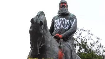 L'image montre une statue équestre d'un homme, probablement un personnage historique, qui porte des graffitis. Le mot "PARDON" est écrit en lettres blanches sur sa robe, tandis que d'autres mots, comme "RACIST", apparaissent en couleur sur le socle. La statue semble être recouverte de peinture, ce qui suggère un acte de protestation ou de critique. En arrière-plan, on peut voir un ciel nuageux et quelques arbres.