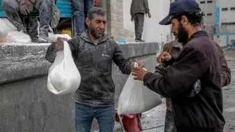 L'image montre des hommes dans un environnement urbain, apparemment en train de distribuer ou de recevoir des sacs. Ils semblent porter des vêtements de travail, certains étant couverts de poussière ou de matériau. L'ambiance suggère une activité liée à l'aide humanitaire ou à une distribution de biens, dans un contexte peut-être de reconstruction ou d'assistance. Les interactions entre les hommes semblent concentrées et sérieuses.