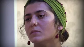 L'image montre une jeune femme avec un regard pensif. Elle porte un foulard vert sur la tête, orné de petites perles. Son visage est en partie encadré par des cheveux bouclés, et elle a un piercing au nez. Elle semble concentrée ou réfléchie, ce qui donne une ambiance contemplative à l'image.