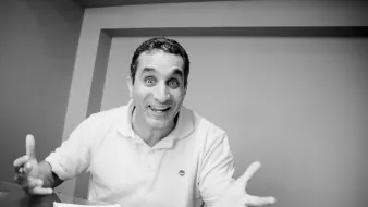 L'image montre un homme assis à une table, avec un air enthousiaste et des gestes expressifs. Il semble parler avec passion, ses bras levés et son visage affichant une grande animation. On peut voir des notes et un petit objet sur la table, suggérant qu'il est en train de discuter ou de partager ses idées. L'image est en noir et blanc, ce qui ajoute une dimension artistique à sa présentation.