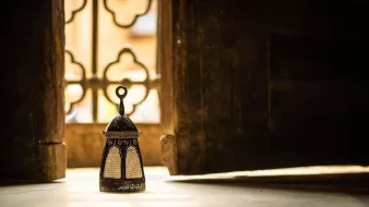 L'image montre une délicate lanterne orientale placée sur le sol, éclairée par une lumière douce, créant une atmosphère chaleureuse. En arrière-plan, on aperçoit un mur avec des motifs décoratifs, et une fenêtre aux formes ornées, laissant transparaître des lumière et ombres intéressantes. La combinaison de la lanterne et de l'environnement évoque une ambiance paisible et méditative.