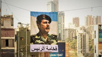 L'image montre une grande bannière suspendue dans une rue urbaine. Sur cette bannière, un portrait d'un homme en uniforme militaire est visible, face à un public. En dessous de l'image, il y a un texte en arabe qui semble faire référence à une autorité ou un leadership. En arrière-plan, on aperçoit des bâtiments modernes et d'autres structures urbaines typiques d'une ville. La lumière est douce, ce qui donne une atmosphère particulière à la scène.