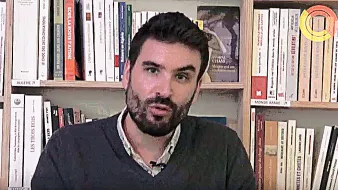 L'image montre un homme assis devant une étagère remplie de livres. Il a une barbe et porte un pull gris. L'expression de son visage semble sérieuse et il semble s'adresser à un auditoire ou à une caméra. L'arrière-plan est constitué de nombreux livres, suggérant un environnement intellectuel ou académique.