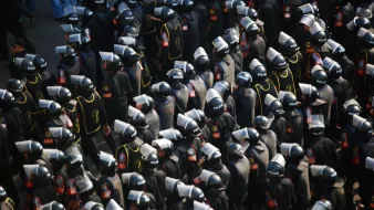 L'image montre un grand groupe de policiers en tenue d'émeute. Ils portent des casques et des boucliers, et sont regroupés de manière organisée. Les uniformes sont sombres, avec des détails visibles tels que des insignes ou des logos indiquant leur affiliation. L'ensemble donne une impression de préparation et de contrôle, évoquant un contexte de maintien de l'ordre lors d'une situation potentiellement tendue.