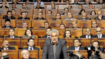L'image montre une séance dans un bâtiment législatif, où une personne (probablement un membre du gouvernement ou un habile orateur) se tient devant un grand groupe de personnes assises. Les membres de l'assemblée, en arrière-plan, semblent concentrés sur le discours prononcé. On peut voir des journalistes avec des caméras, suggérant que cet événement est couvert par les médias. L'atmosphère est formelle et sérieuse, typique de débats ou de discussions politiques.