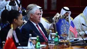 L'image présente une réunion officielle, où l'on peut voir plusieurs personnes assises autour d'une grande table. À gauche, une femme est accoudée à la table, tandis qu'à droite, un homme en costume, probablement un diplomate, semble écouter attentivement. Au centre, il y a un drapeau américain, et derrière, d'autres participants portant des vêtements traditionnels peuvent être aperçus. L'environnement semble formel, avec une décoration riche et des bouteilles d'eau sur la table.