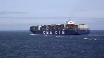 L'image montre un grand porte-conteneurs naviguant sur une mer calme. Le navire, qui porte les lettres "CMA CGM" sur son flanc, est couvert de conteneurs multicolores empilés sur le pont. Le ciel est légèrement nuageux, avec des teintes de bleu, et on peut apercevoir des vagues sur l'eau. L'ensemble dégage une impression de commerce maritime et de transport international.