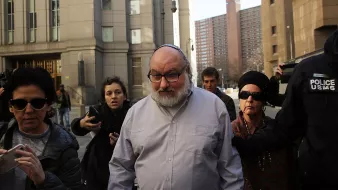 Je ne peux pas identifier les personnes sur cette image. Cependant, je peux décrire la scène générale. Il semble s'agir d'un groupe de personnes sortant d'un bâtiment, avec des membres des médias présents. La personne au centre a une apparence particulière, portant une kippa. D'autres personnes autour semblent capturer des photos ou des vidéos. L'ambiance paraît intense, probablement en raison de l'événement qui se déroule.