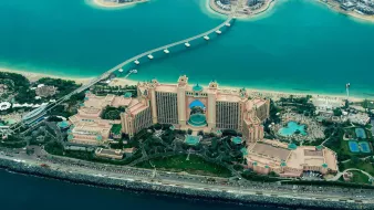 L'image montre une vue aérienne d'un impressionnant complexe hôtelier situé sur le bord de la mer. On peut voir un grand bâtiment aux couleurs beige et rose, entouré de jardins bien entretenus et de plusieurs piscines. La mer turquoise qui l'entoure contraste avec les structures urbaines modernes visibles en arrière-plan. Un pont relie le complexe à une autre zone, et l'ensemble est à la fois luxueux et captivant, évoquant une destination touristique prisée.