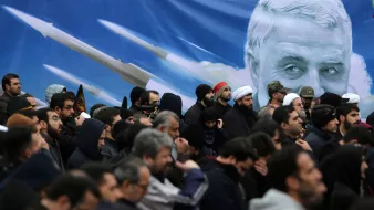 L'image montre une foule de personnes rassemblées, vraisemblablement lors d'un événement public. Les individus portent des vêtements sombres et certains semblent tenir des drapeaux ou des bannières. En arrière-plan, il y a une grande illustration d'un visage avec des images de missiles, ce qui pourrait indiquer un contexte politique ou militaire. L'ambiance générale semble sérieuse et solennelle.