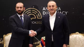 L'image montre deux hommes se serrant la main, probablement en signe de coopération ou de négociation. Ils sont vêtus de costumes formels. L'arrière-plan présente le logo d'un événement lié à la diplomatie, avec la mention "Ankara Diplomacy 2023". L'ambiance semble être celle d'une rencontre officielle.