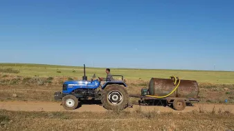 L'image montre un homme conduisant un tracteur bleu sur une route de terre. Il tire une remorque avec un grand réservoir métallique. À l'arrière-plan, on peut voir des champs verts sous un ciel dégagé. La scène évoque une ambiance rurale et agricole.
