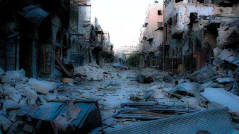 L'image montre une rue dévastée, probablement après un conflit ou une catastrophe. On peut voir des bâtiments endommagés avec des murs effondrés et des débris éparpillés sur le sol. Le contexte semble être celui d'une ville abandonnée, avec une atmosphère sombre et désolée. Les traces de destruction sont évidentes, illustrant les ravages subis par l'environnement urbain.