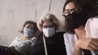 Sur l'image, on peut voir trois personnes assises côte à côte, chacune portant un masque. La première personne à gauche a les cheveux bouclés et porte un haut clair. La personne au milieu a des cheveux gris et porte des lunettes ainsi qu'un haut noir. La dernière personne à droite a des cheveux bouclés et porte également un haut clair. Elles semblent se détendre ensemble, peut-être dans un environnement public ou en extérieur. L'ambiance est informelle et conviviale.